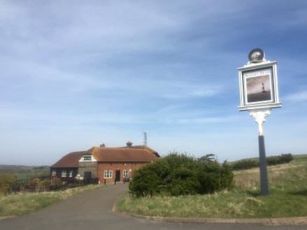 A brick building with a stone doorway and thatched roof which contains the Beachy Head Pub/Inn/Shop/Museum. A sign with a painted picture of the Beachy Head Lighthouse on the lefthand side.